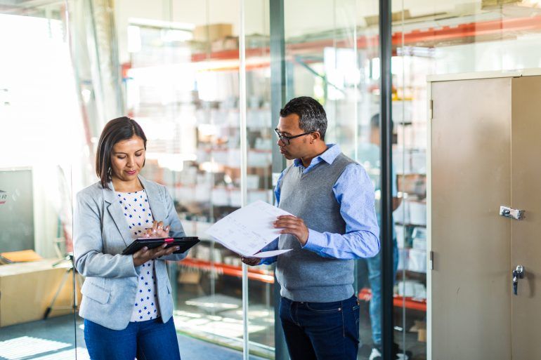 Two business professionals reviewing inventory documents for MCA 2 million dollar loans on a tablet inside a warehouse or logistics facility.