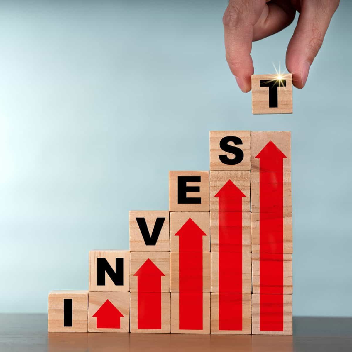 Wooden blocks stacked upward spelling “INVEST” with red growth arrows and a hand placing the final block on top, symbolizing business financing and growth in a comparison of 8fig vs Swish Funding.