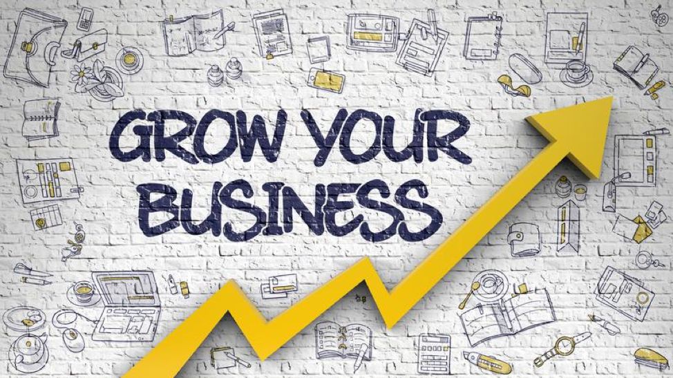 A white brick wall background with hand-drawn business and technology icons in black and yellow. In the center, bold navy blue text says "GROW YOUR BUSINESS." A large yellow upward arrow overlays the text, symbolizing growth and progress.